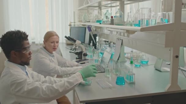Waist up van multiraciale paar chemici in lab jassen praten terwijl het maken van experimenten in glaswerk, samen te werken in het lab - Video