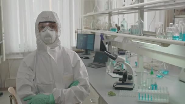 Medium portret van blanke vrouwelijke wetenschapper in veiligheidsbril, ademhalingsmasker en beschermende bekleding poseren met de handen gevouwen in het laboratorium - Video