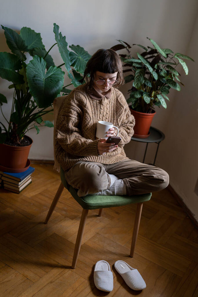Інтроверт підліток проводить час один вдома, читає новини в Інтернеті, прокручує соціальні мережі, обмінюється повідомленнями в смартфоні. Дівчина сидить у затишній кімнаті з кімнатами, які п'ють чашку чаю - Фото, зображення