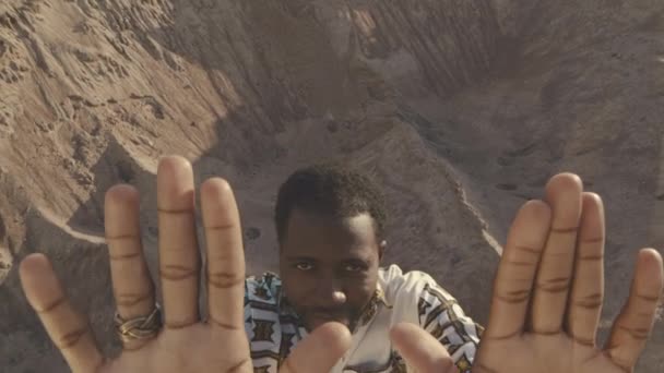 Zoom out πλάνο της ισχυρής Αφρικής Αμερικανός νεαρός άνδρας σε περίτεχνα jumpsuit σπρώχνοντας κάμερα μακριά με τα χέρια του και κοιτάζοντας απευθείας σε σας, ενώ στέκεται μακριά σε αμμόλοφους - Πλάνα, βίντεο