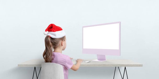 Χριστουγεννιάτικο καπέλο λειτουργεί σε οθόνη υπολογιστή mockup σε ένα επίπεδο γραφείο. Εποχιακή απομακρυσμένη εργασία με πνεύμα διακοπών - Φωτογραφία, εικόνα