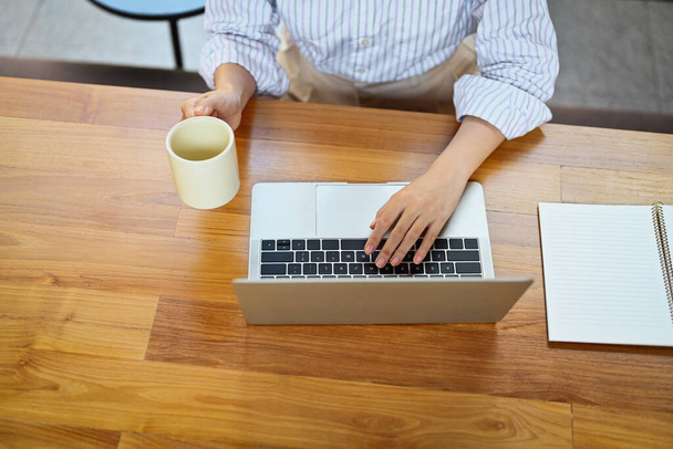 Над просмотром женщина-офисный работник держит чашку кофе и работает на ноутбуке. - Фото, изображение