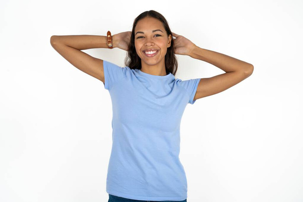 Ικανοποιημένη νεαρή όμορφη ισπανόφωνη γυναίκα φορώντας μπλε μπλουζάκι πάνω από το λευκό φόντο κρατήστε τα χέρια πίσω από το κεφάλι χαλαρώνοντας - Φωτογραφία, εικόνα