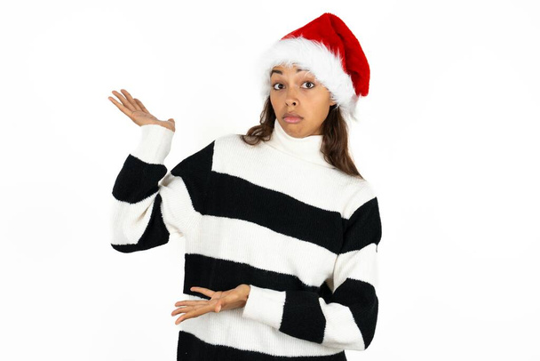 Молода красива жінка в смугастому светрі і капелюсі Санта-Клауса, спрямовані в сторону обох рук, показуючи щось дивне і кажучи: "Я не знаю, що це. Концепція реклами. - Фото, зображення