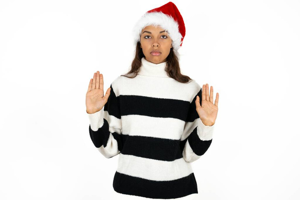Serious Young beautiful woman wearing striped sweater and a santa claus hat pulls palms towards camera, makes stop gesture, asks to control your emotions and not be nervous - Photo, Image