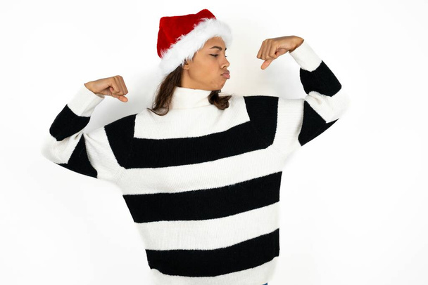 Молодая красивая женщина в полосатом свитере и шляпе Санта Клауса, показывающая мышцы рук, улыбающиеся с гордостью. Фитнес-концепция. - Фото, изображение