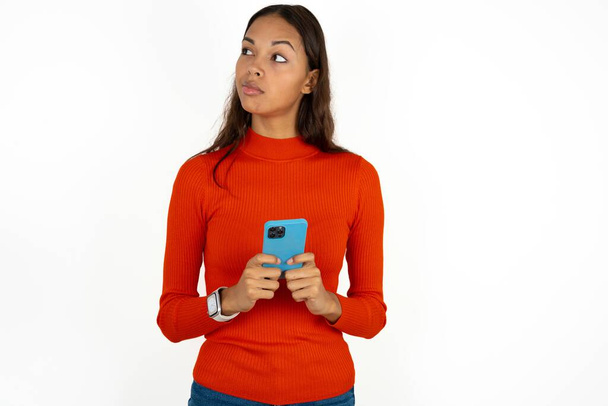 joven hispano mujer usando rojo cuello alto sobre blanco fondo sostiene teléfono manos lee buena juventud noticias mirada vacío espacio anuncio - Foto, imagen