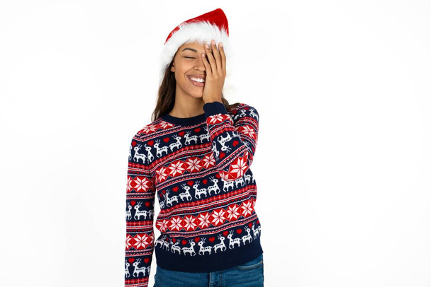 красивая латиноамериканка в трикотажном свитере и шляпе Санта Клауса на белом фоне делает лицо ладонью и широко улыбается, хихикает положительно слышит смешные шутки позы - Фото, изображение