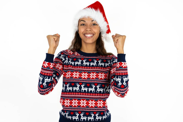 Schockiert ekstatisch schöne hispanische Frau in Strickpullover und Weihnachtsmann Hut über weißem Hintergrund gewinnen Glück Lotterie heben die Hände schreien ja - Foto, Bild