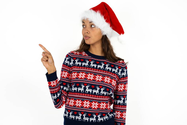 beautiful hispanic woman wearing knitted sweater and santa claus hat over white background points at copy space and advertises something, advices best price. - Photo, Image