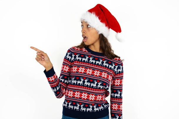 Stunned beautiful hispanic woman wearing knitted sweater and santa claus hat over white background with greatly surprised expression points away on copy space, indicates something - Photo, Image