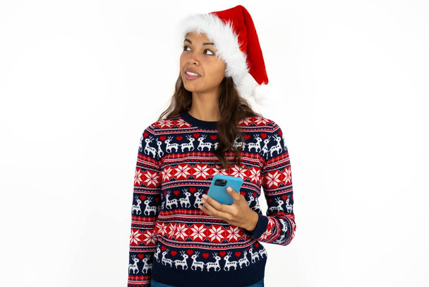 gyönyörű spanyol nő visel kötött pulóver és Santa Claus kalap fehér háttér tartja mobiltelefon használ nagy sebességű internet és a szociális hálózatok online kommunikáció. Modern technológiák koncepciója - Fotó, kép