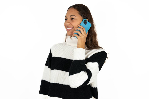 ホワイトバックグラウンドのトークでストライプセーターを着用している若い美しい女性は,携帯電話の笑顔良い気分を話す - 写真・画像