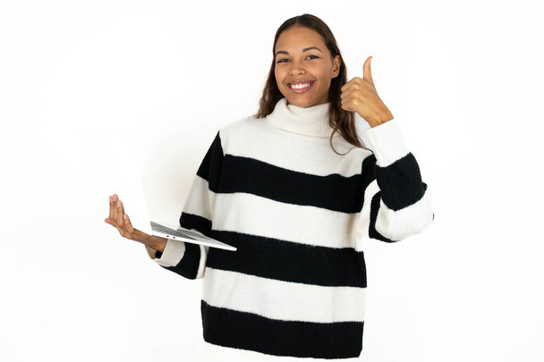 Bella attraente giovane bella donna che indossa maglione a righe su sfondo bianco tenere netbook mano dito mostrare pollice in su - Foto, immagini