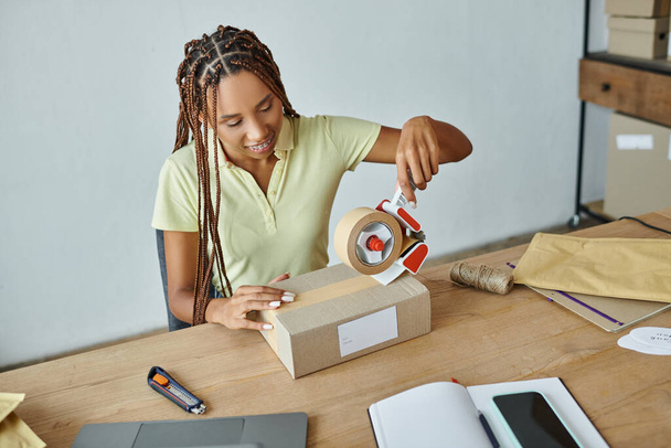 jolly young african american female retailer using tape on cardboard box, delivery concept - Photo, Image