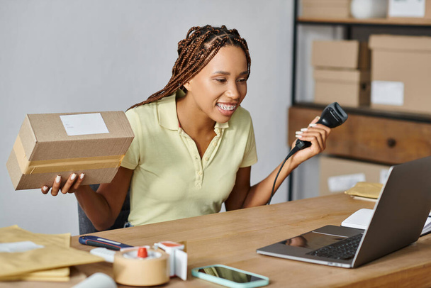 alegre africano americano comerciante femenino mirando el ordenador portátil mientras escanea la caja, concepto de entrega - Foto, imagen