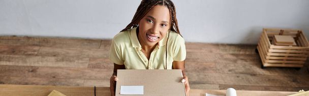 vidám csinos afro-amerikai eladó holding box és mosolyog a kamera, szállítási koncepció, banner - Fotó, kép