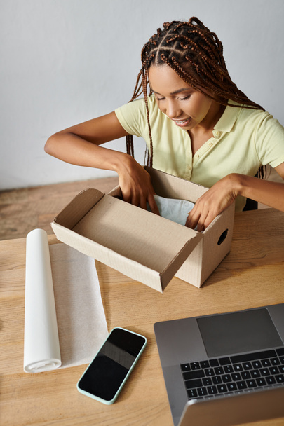 radosna młoda Afroamerykanka pakująca pudełko i uśmiechająca się szczęśliwie pracując ciężko, dostawa - Zdjęcie, obraz