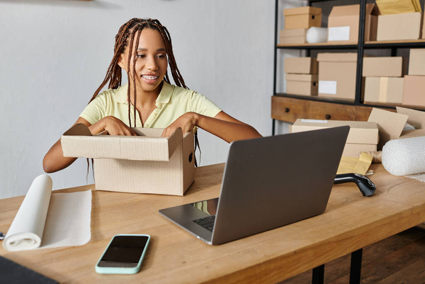 vidám fiatal afro-amerikai női kereskedő csomagolás doboz és nézi laptop, szállítás - Fotó, kép