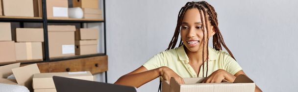 gioiosa attraente scatola di imballaggio donna africana americana venditore e guardando laptop, consegna, banner - Foto, immagini