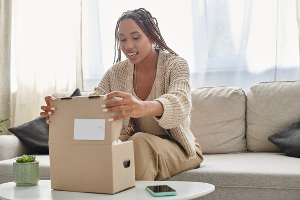 fröhliche junge afrikanisch-amerikanische Frau in kuscheliger Homewear sitzt auf dem Sofa und öffnet ihr Paket - Foto, Bild