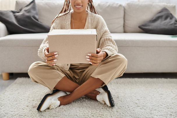 vista cortada da mulher americana africana alegre com aparelho sentado no chão e segurando parcela - Foto, Imagem