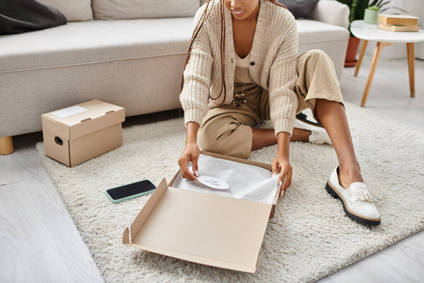 cropped view of jolly young african american woman sitting on floor and opening cardboard parcel - Photo, Image