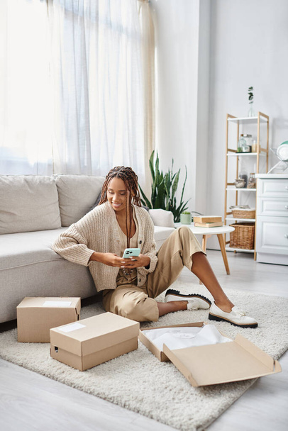 jolly appealing african american woman in homewear holding phone and looking at parcels on floor - Photo, Image