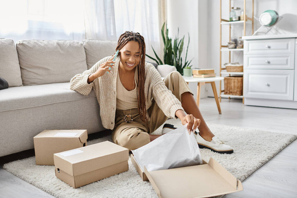 attractive jolly african american woman talking by phone while sitting on floor next to her parcel - Photo, Image