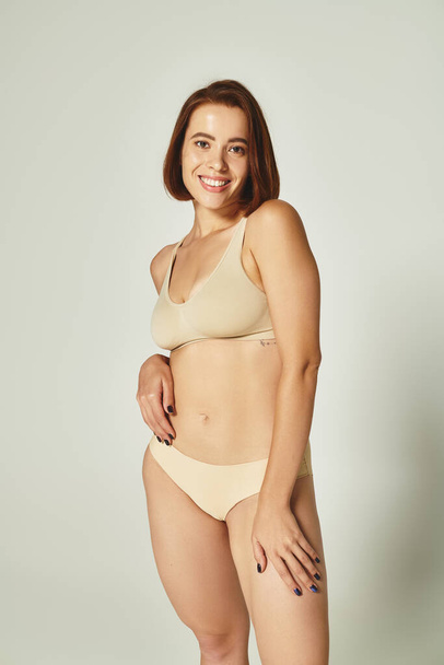 lichaam positieve, vrolijke jonge vrouw met kort haar staan in beige ondergoed op grijze achtergrond - Foto, afbeelding