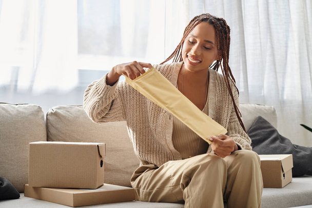 alegre joven afroamericana mujer apertura post paquete mientras está sentado en sofá rodeado de paquetes - Foto, Imagen