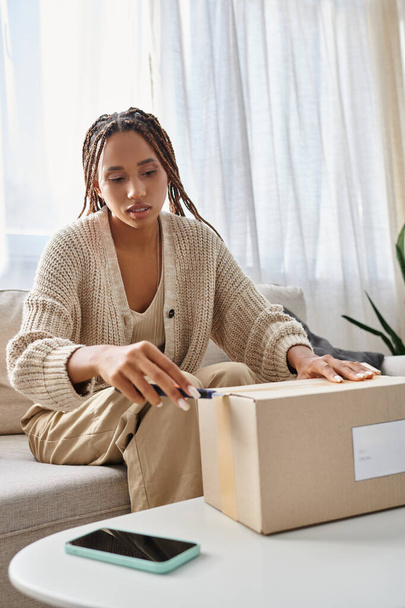 young attractive african american woman in homewear using stationery knife to open her parcel - Photo, Image