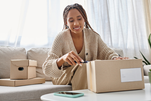 jolly african american woman in casual attire using stationery knife to open cardboard box - 写真・画像