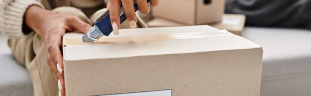 cropped view of african american woman using stationery knife to open cardboard parcel, banner - Photo, Image
