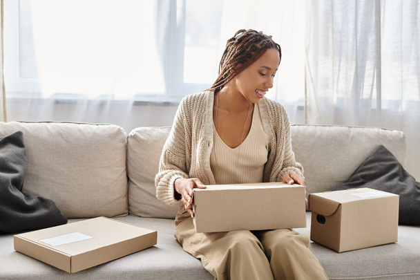 good looking jolly african american woman sitting on sofa and preparing to open cardboard box - Photo, Image