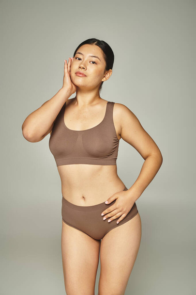 Kahverengi iç çamaşırlarıyla gri arka planda poz veren esmer Asyalı çekici bir genç kadın. - Fotoğraf, Görsel