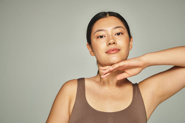 bonita mujer asiática en marrón superior tocando su cara sobre fondo gris, cuidado de la piel y belleza - Foto, Imagen