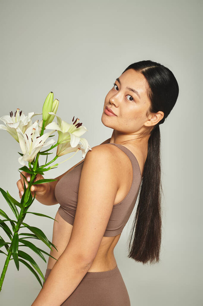 brunetta donna asiatica in biancheria intima pastello che tiene fiori e in piedi su sfondo grigio, gigli - Foto, immagini