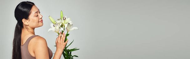 elégedett fiatal ázsiai nő pasztell fehérnemű nézi virágok szürke háttér, banner - Fotó, kép