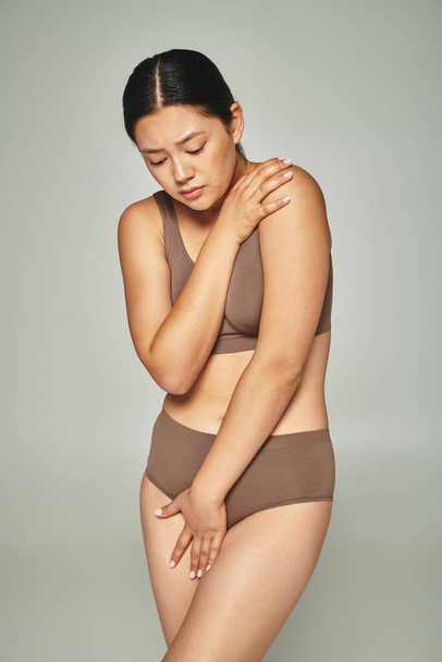 грустная азиатская женщина в нижнем белье покрывает тело, обнимая себя на сером фоне, тело стыдится - Фото, изображение