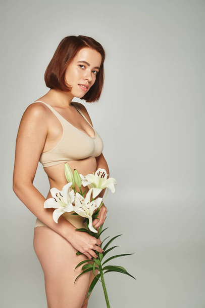 charmante vrouw met kort haar houden bloemen en poseren in ondergoed op grijze achtergrond, lelies - Foto, afbeelding