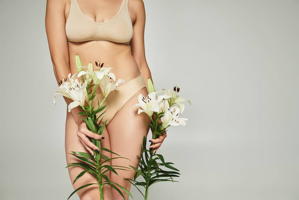 kobieta w beżowej bieliźnie trzyma białe lilie i stoi na szarym tle, widok przycięty - Zdjęcie, obraz