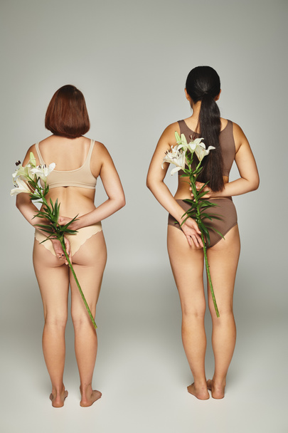 widok z tyłu dwie kobiety w beżowej bieliźnie stojące z białymi liliami w dłoniach na szarym tle - Zdjęcie, obraz