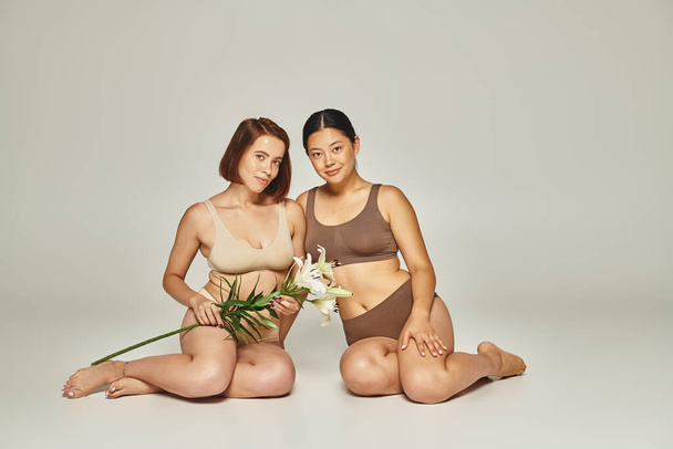 jonge multiculturele vrouwen in ondergoed zitten samen met bloemen op grijze achtergrond, lelies - Foto, afbeelding