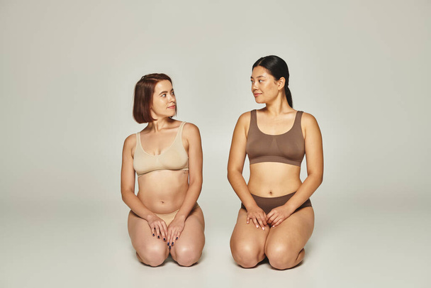 two diverse women in beige and brown underwear sitting together on grey background, body positive - Photo, Image