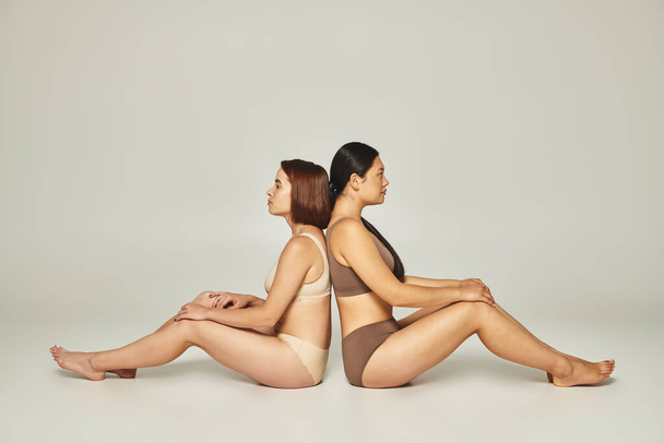 duas amigas diversas em roupa interior bege e marrom sentadas de costas para trás no fundo cinza - Foto, Imagem