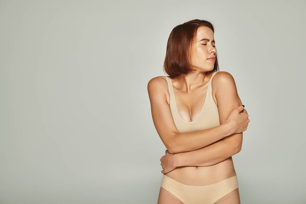 bezorgde jonge vrouw in beige ondergoed kijken over de schouder op grijze achtergrond, kwetsbaarheid - Foto, afbeelding