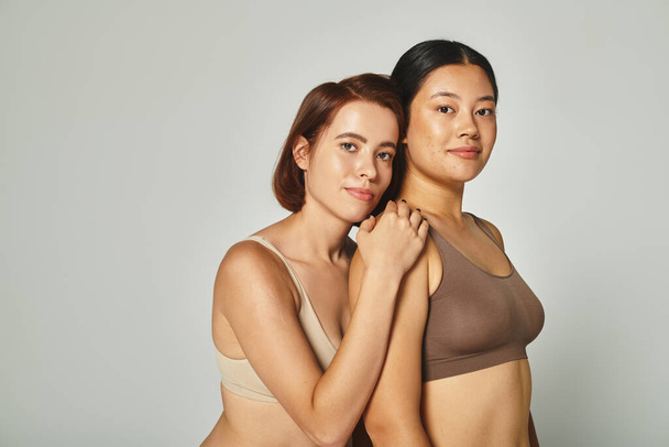 mladé multikulturní ženy ve spodním prádle objímající a dívající se na kameru na šedém pozadí - Fotografie, Obrázek