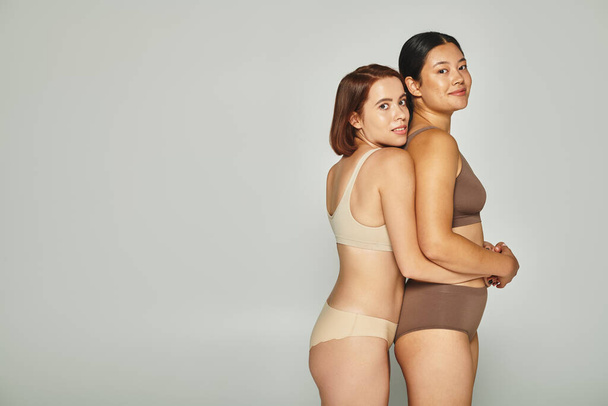 young multicultural women in pastel underwear embracing and looking at camera on grey background - Photo, Image