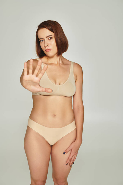 Nuori nainen beige alusvaatteet ele ja osoittaa lopettaa harmaalla pohjalla, kehon häpeä - Valokuva, kuva